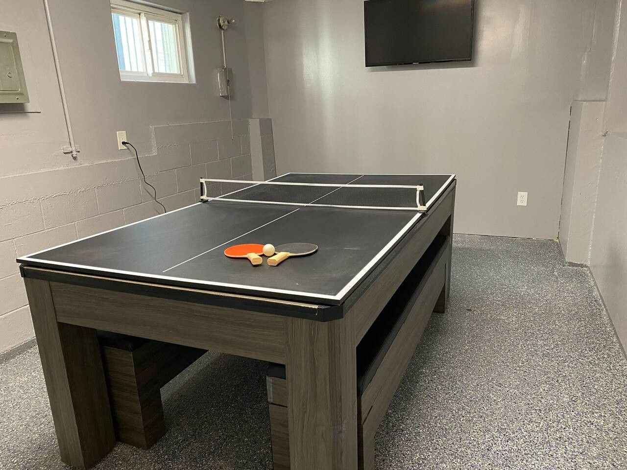 A ping pong table with paddles and balls in a room with a tiled floor and a television mounted on the wall.