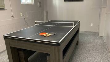 A ping pong table with paddles and balls in a room with a tiled floor and a television mounted on the wall.