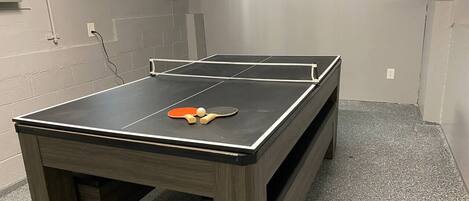 A ping pong table with paddles and balls in a room with a tiled floor and a television mounted on the wall.