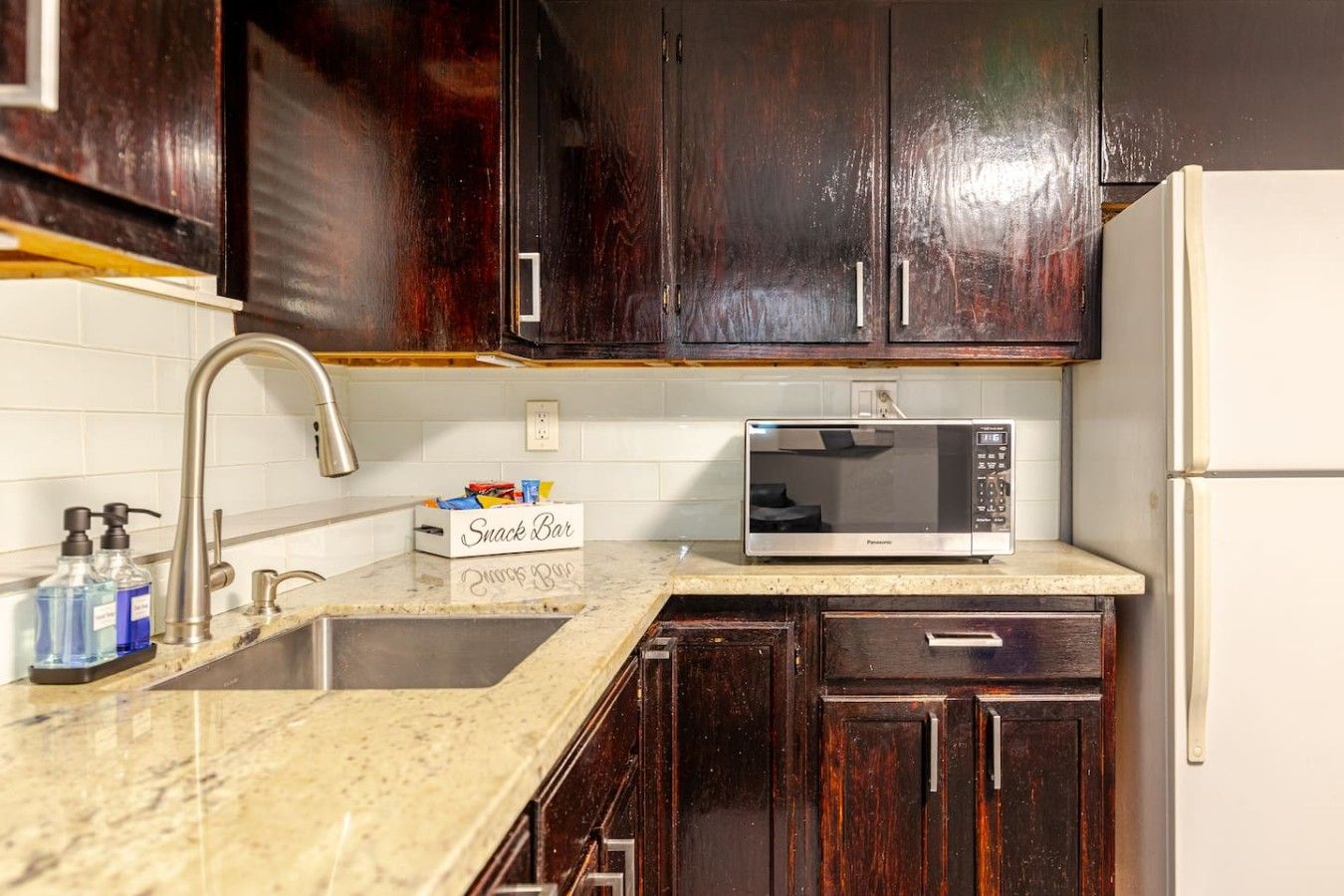 A kitchen with dark wooden cabinets, a white refrigerator, a stainless steel sink, and a microwave on the countertop.
