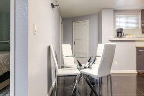 Un espacio habitable compacto con comedor que incluye una mesa de vidrio y sillas blancas, una kitchenette con cafetera y una puerta que da a otra habitación.