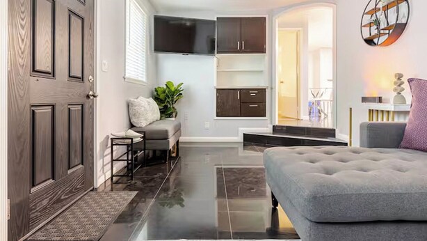 Una sala de estar moderna con un sofá gris, una mesita y un banco gris. Hay una puerta a la izquierda y una escalera que lleva a otra habitación.