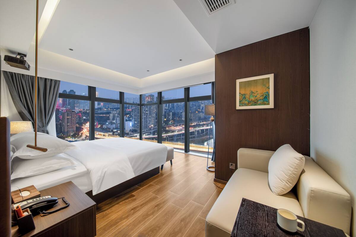 Une chambre d’hôtel moderne dotée d’un grand lit, d’un canapé, d’une table basse, d’un bureau avec un téléphone et offrant une vue sur la ville grâce à de grandes fenêtres.