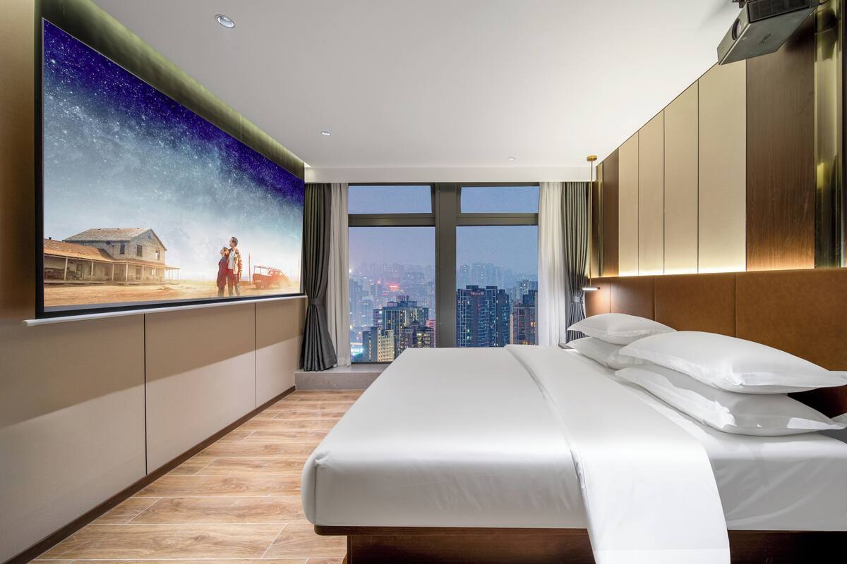 Une chambre d’hôtel moderne avec un grand lit, une vue sur la ville et un téléviseur fixé au mur qui diffuse une scène de film.