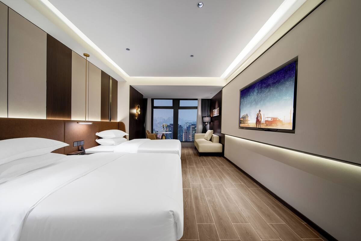 Une chambre d’hôtel avec deux lits, un canapé, une grande fenêtre donnant sur la ville et un tableau au mur.