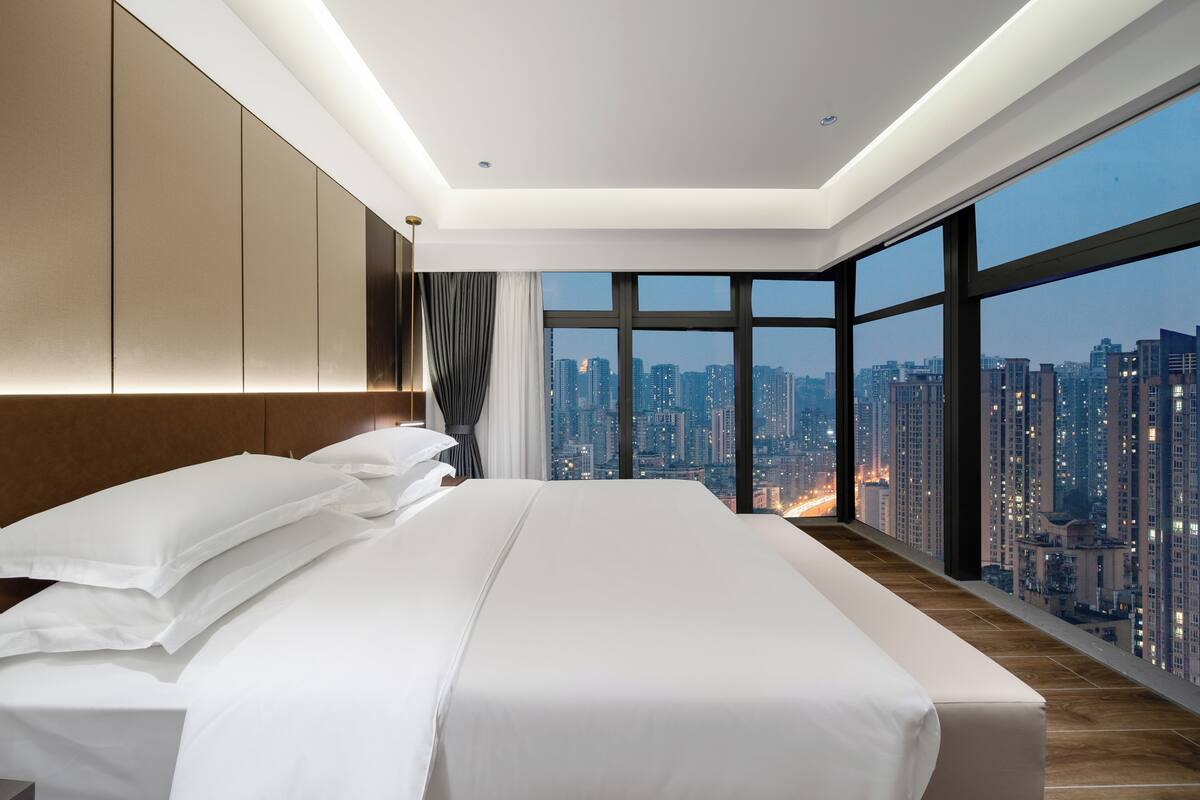 Une chambre d’hôtel moderne avec un grand lit, une vue sur les toits de la ville et un plafond haut.