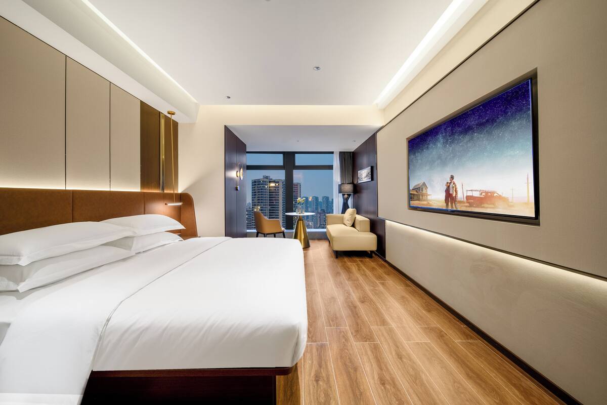 Une chambre d’hôtel avec un grand lit, une vue sur la ville, un tableau au mur et une petite table avec une chaise.