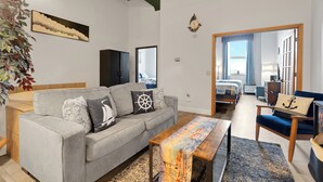 Un moderno salón con un sofá gris, una mesa de centro de madera y un espejo grande que refleja la habitación. Se ve una planta a la izquierda y, al fondo, una cama con cabecera azul.