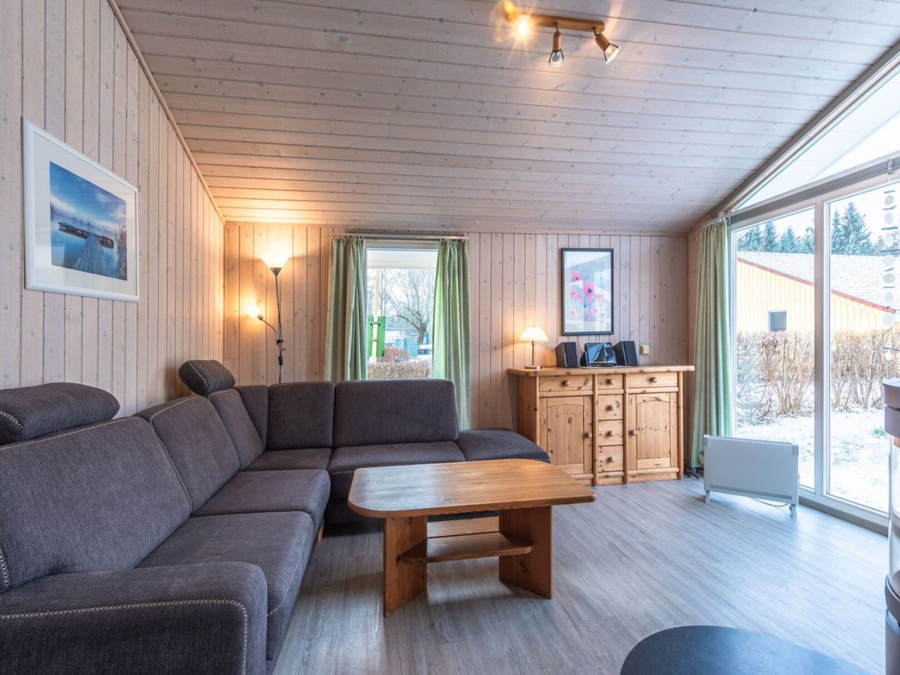 A cozy cabin-style room with a sectional sofa, a wooden coffee table, and a wooden cabinet. There are two windows with curtains, a wall-mounted lamp, and framed pictures.