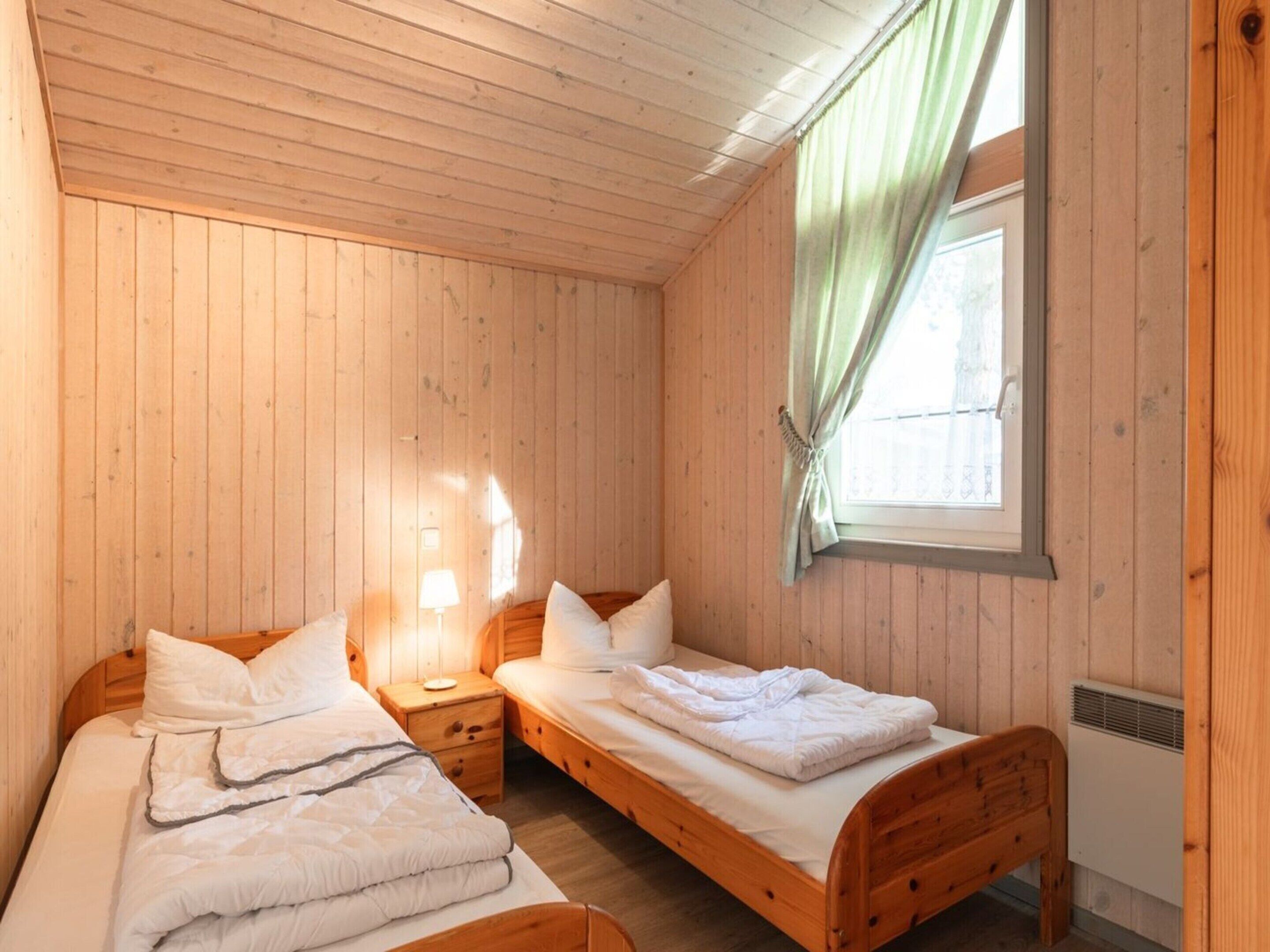 Una habitación pequeña y acogedora con paredes de madera y una ventana con cortinas. Hay dos camas individuales con estructura de madera y ropa de cama blanca. Junto a cada cama, una mesita de noche con una lámpara.