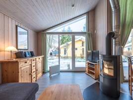 L’intérieur chaleureux d’un chalet avec des murs en bois, une grande porte-fenêtre coulissante donnant sur une cour enneigée, une cheminée intégrée et un téléviseur posé sur un meuble en bois.