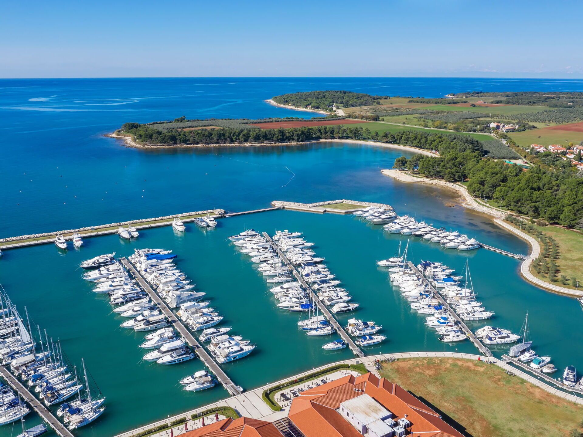 An aerial view of a marina with numerous boats docked, surrounded by greenery and a clear blue sea.