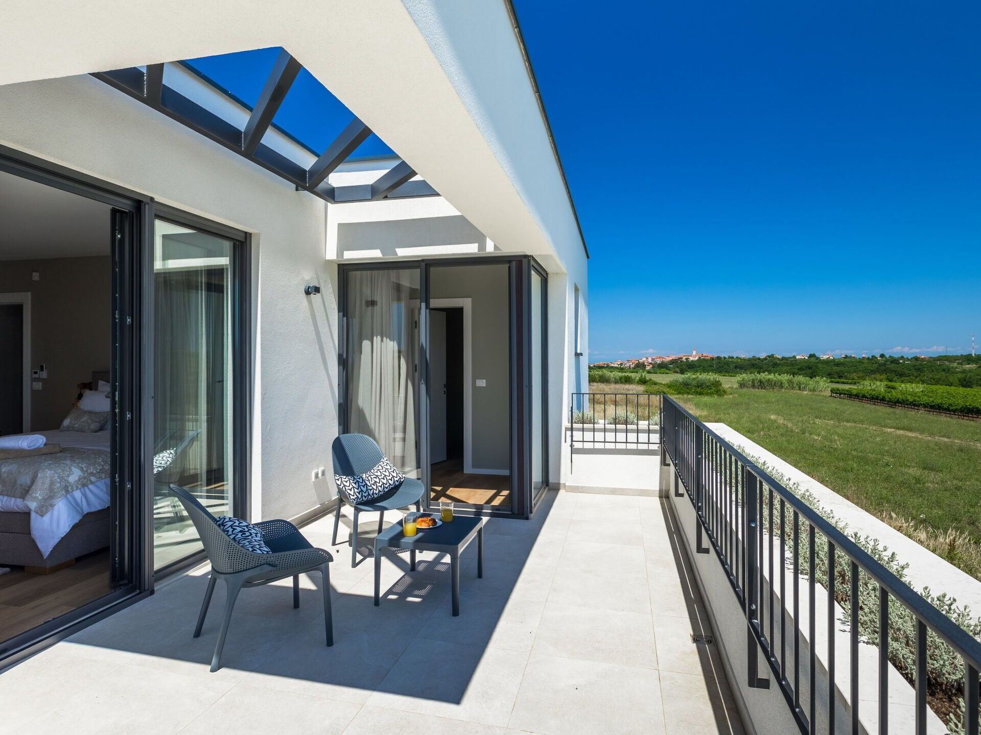 Een balkon met een glazen deur naar een slaapkamer, een tafel met een stoel, en uitzicht op een groen landschap.
