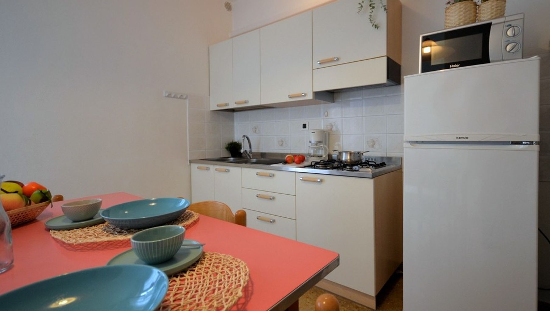 A compact kitchen with white cabinets, a stove, a microwave, and a refrigerator. There is a table with bowls and a basket of fruit.