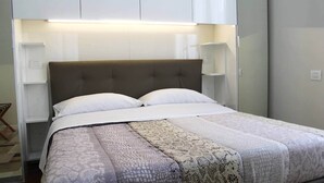 A neatly made bed with a patterned bedspread and pillows, flanked by white bedside tables with lamps, in a room with a simple, clean design.