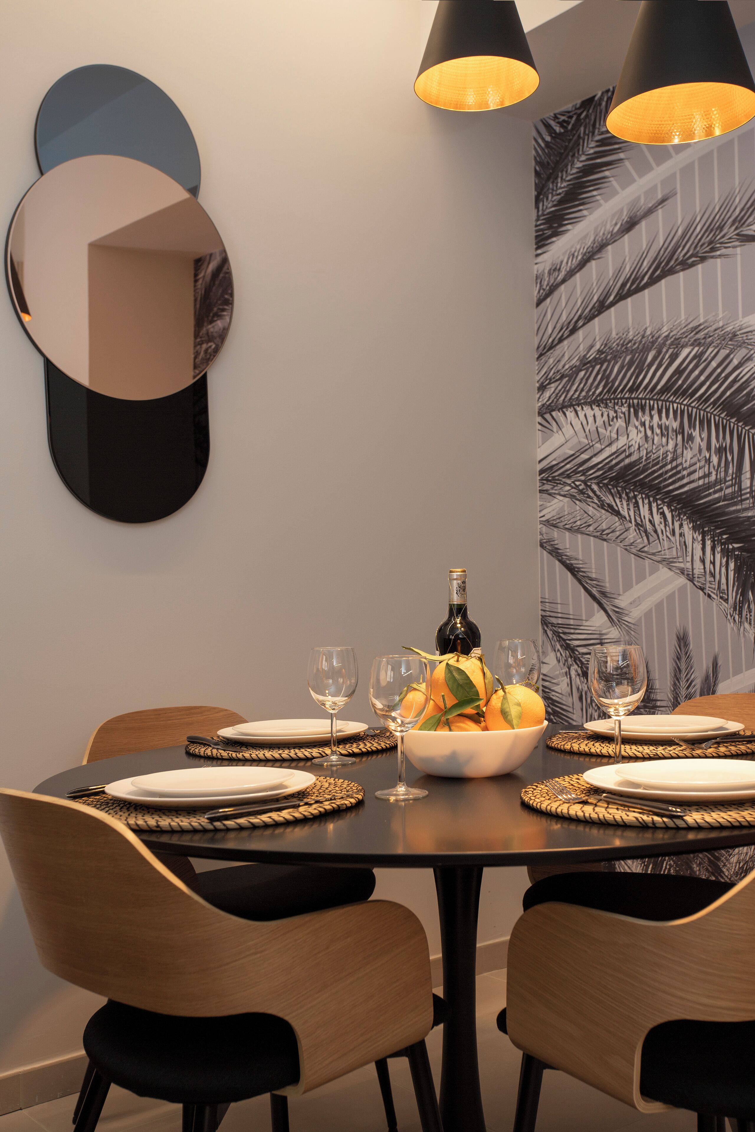 Una mesa de comedor dispuesta con platos, vasos y un frutero, rodeada de sillas.