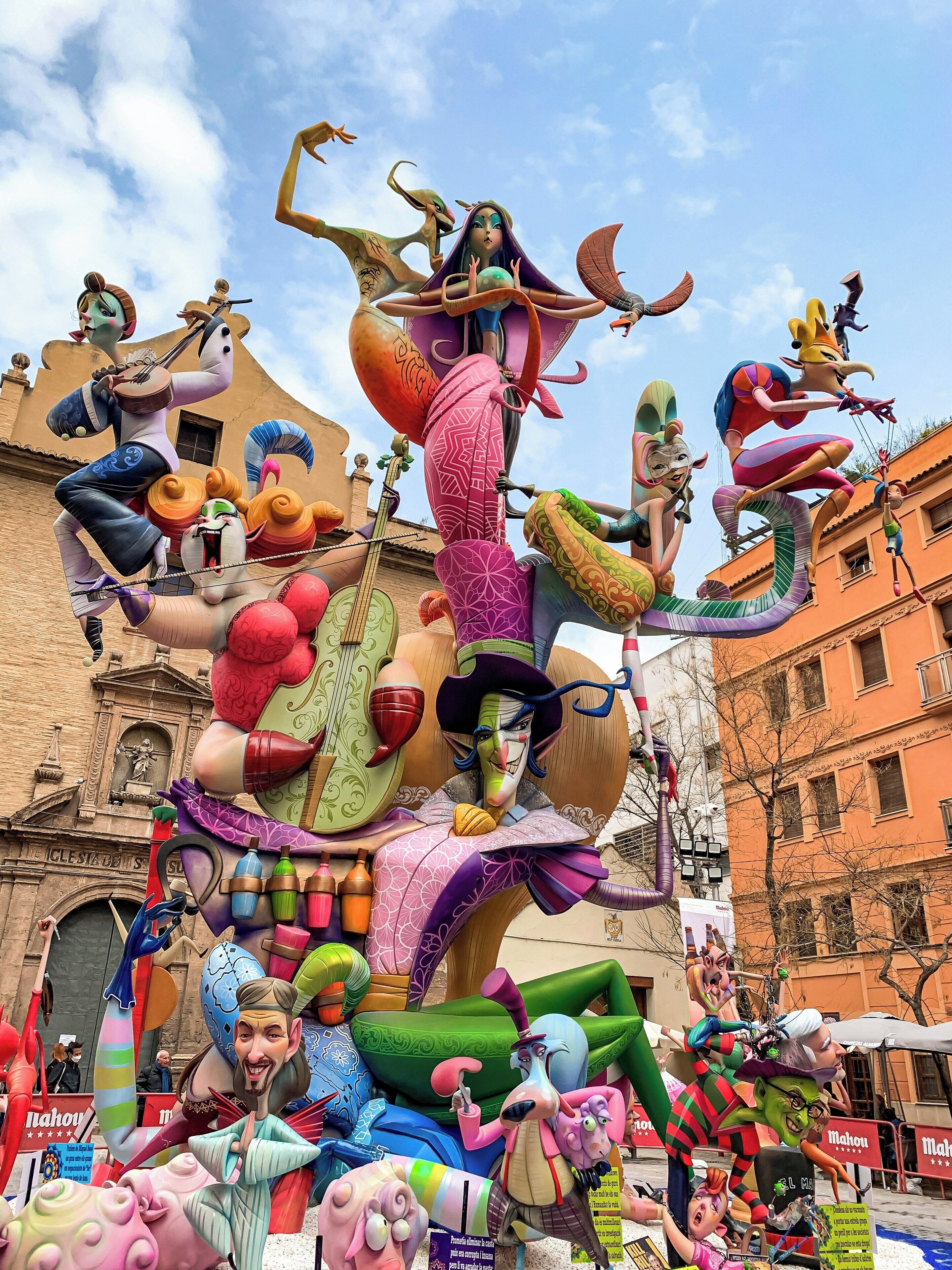 Une grande sculpture colorée, représentant divers personnages et objets, placée devant un bâtiment à l’architecture classique.
