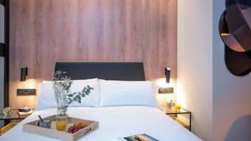 Una cama bien hecha con ropa de cama blanca, una bandeja con alimentos para el desayuno, un libro y un florero con flores.