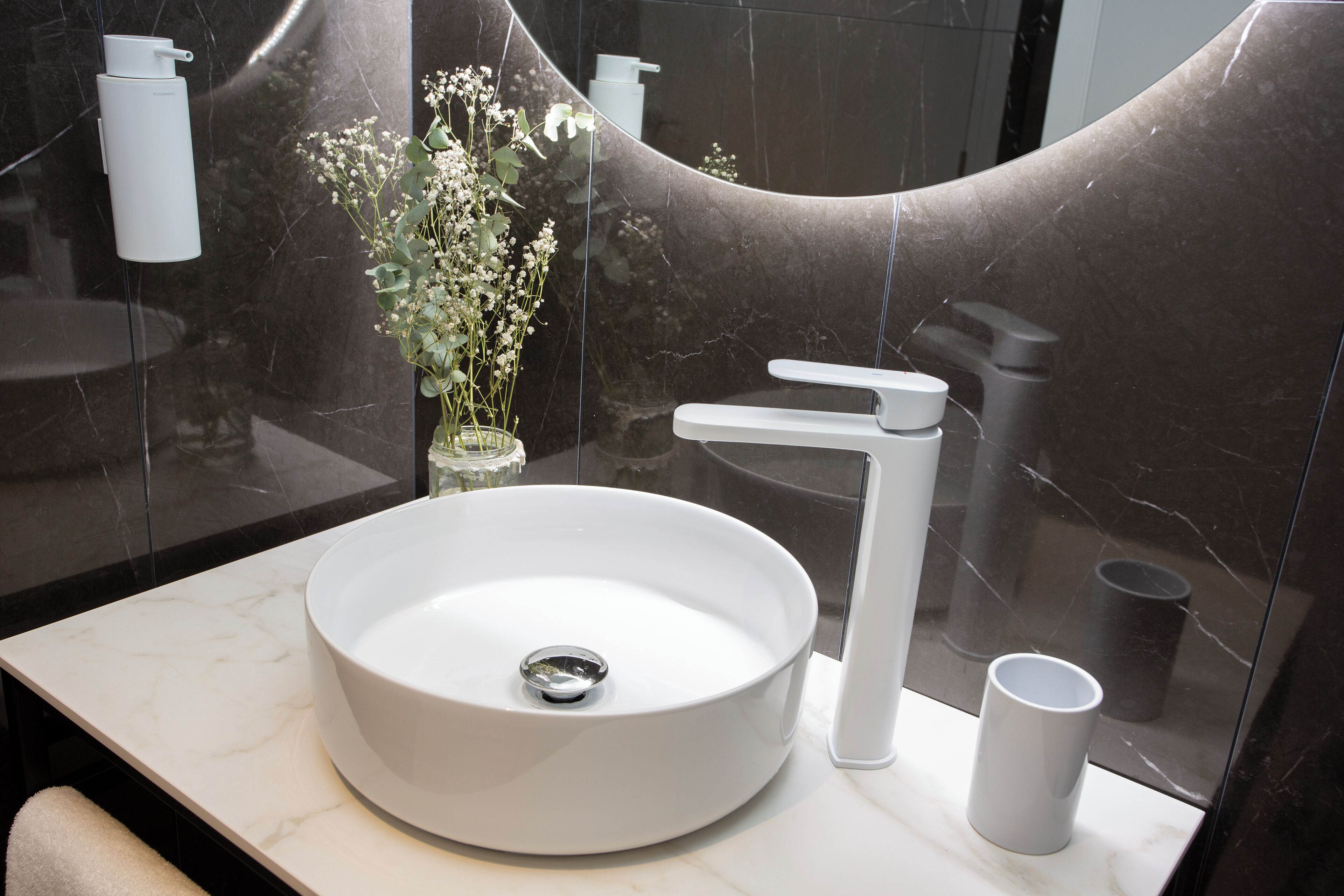 Un baño moderno con un lavabo blanco redondo, un solo grifo y un pequeño jarrón con flores.