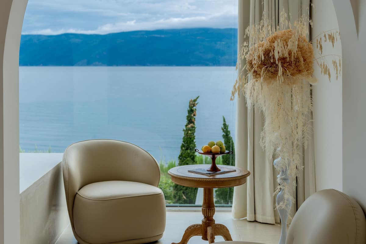 A room with a view of a lake and mountains, featuring two leather chairs and a small table with a decorative item.