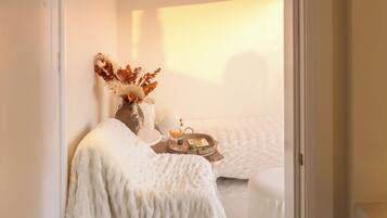 Un dormitorio acogedor con una cama blanca, una mesita de noche con lámpara y un jarrón con flores secas.