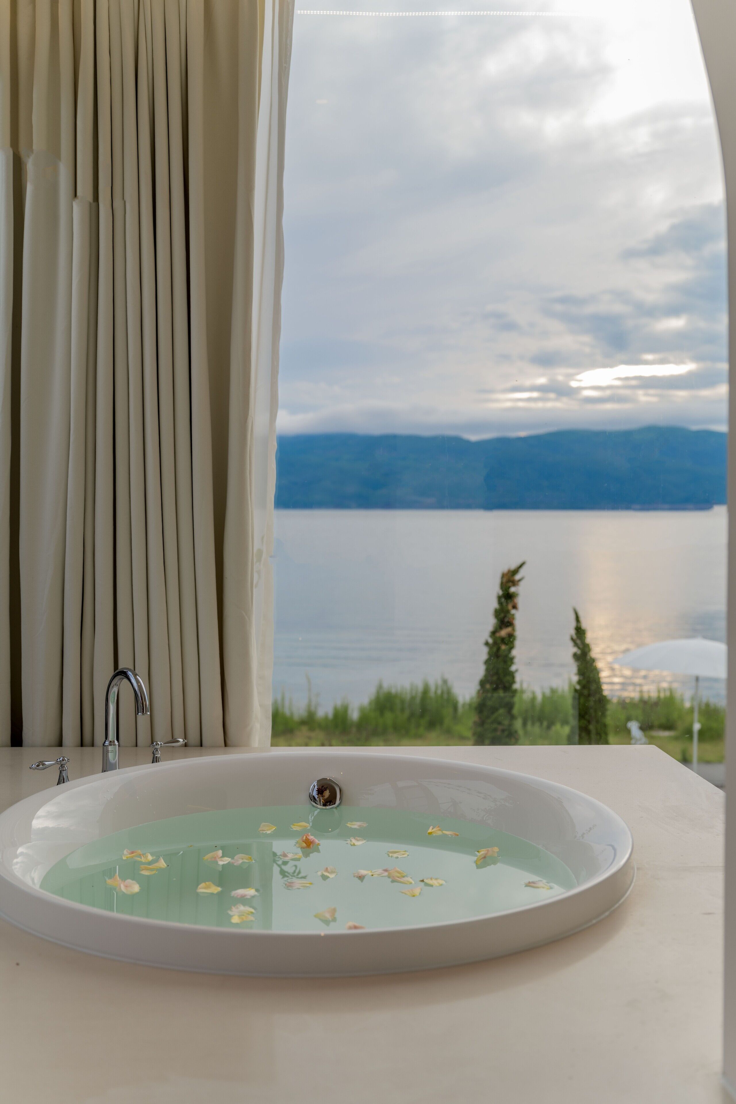 A bathroom with a bathtub filled with water and flower petals, a window with curtains, and a view of a lake and mountains.