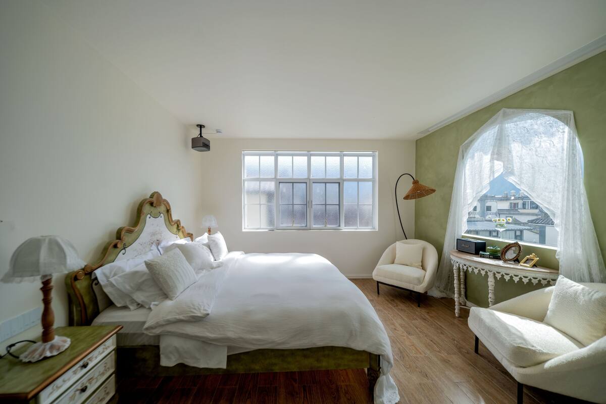 A bedroom with a large bed, a chair, a small table, and a window with a view of buildings.