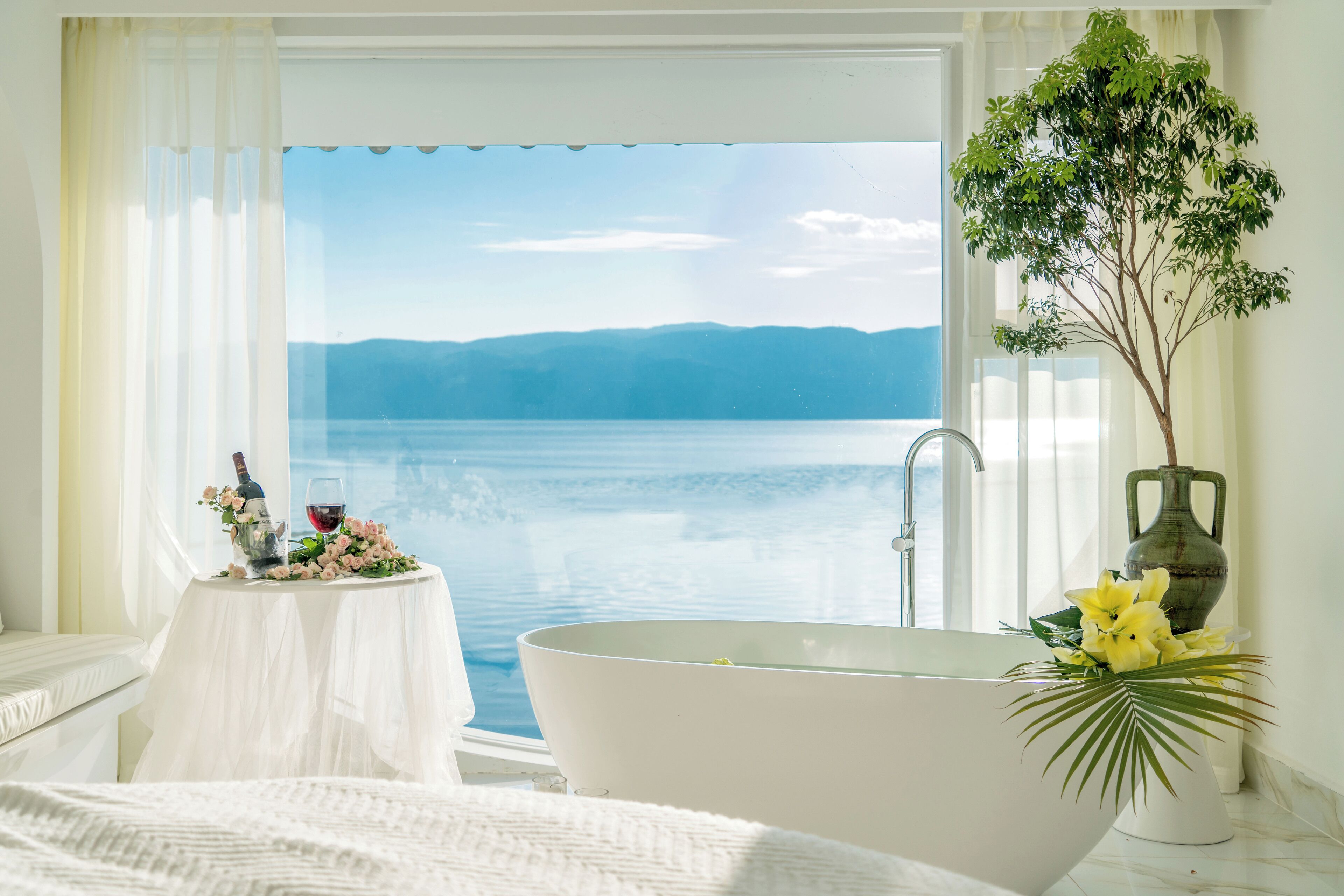 A bathroom with a bathtub, a small table with flowers and a bottle, and a view of a lake and mountains.