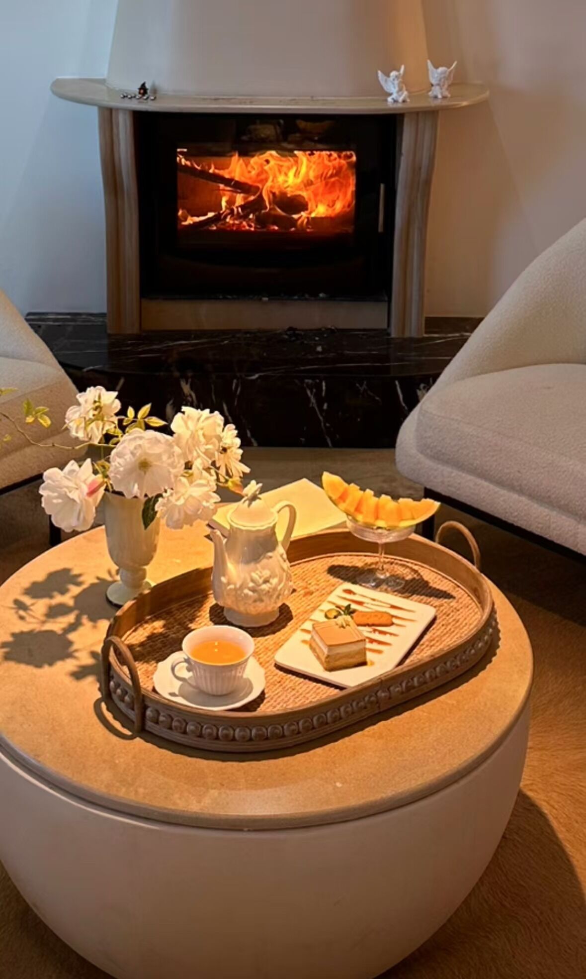 Ein gemütliches Wohnzimmer mit Kamin, einem runden Couchtisch mit Blumenarrangements, einer Teekanne und einer Desserttablett.