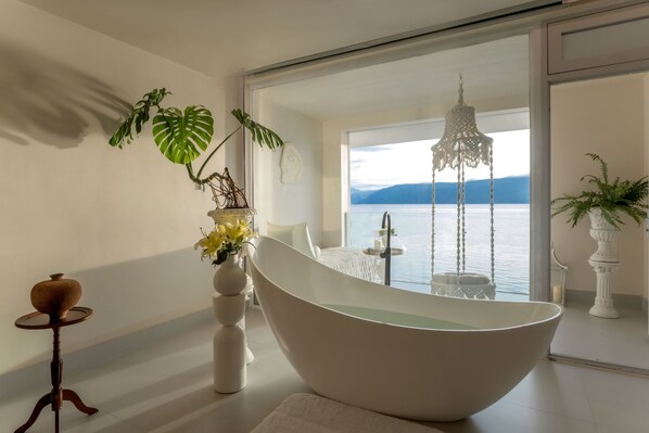 A modern bathroom with a freestanding bathtub, a large window with a view of the sea, and a chandelier.