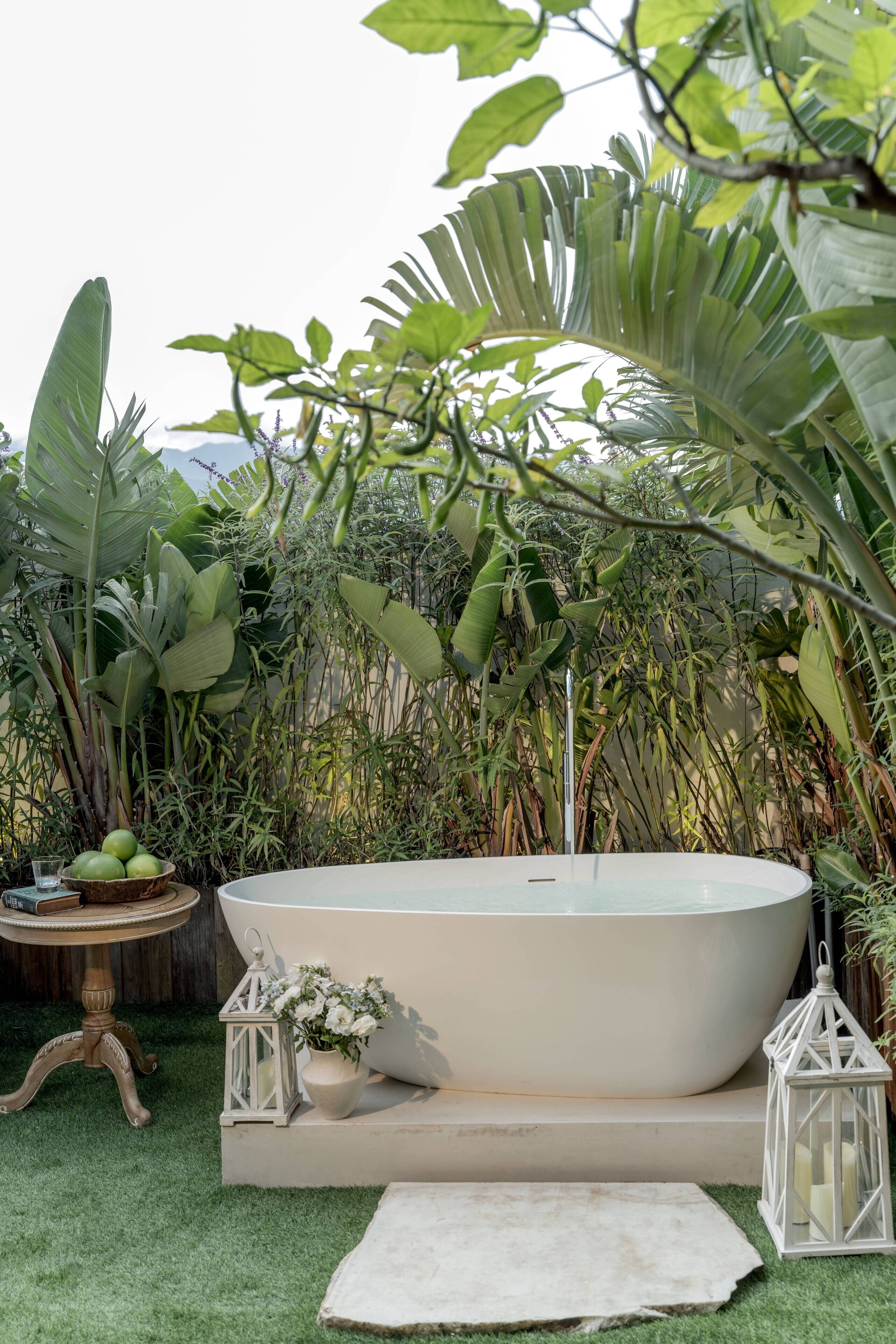 Un área de spa con una bañera grande, rodeada de exuberante vegetación y una pequeña mesa con velas y flores.