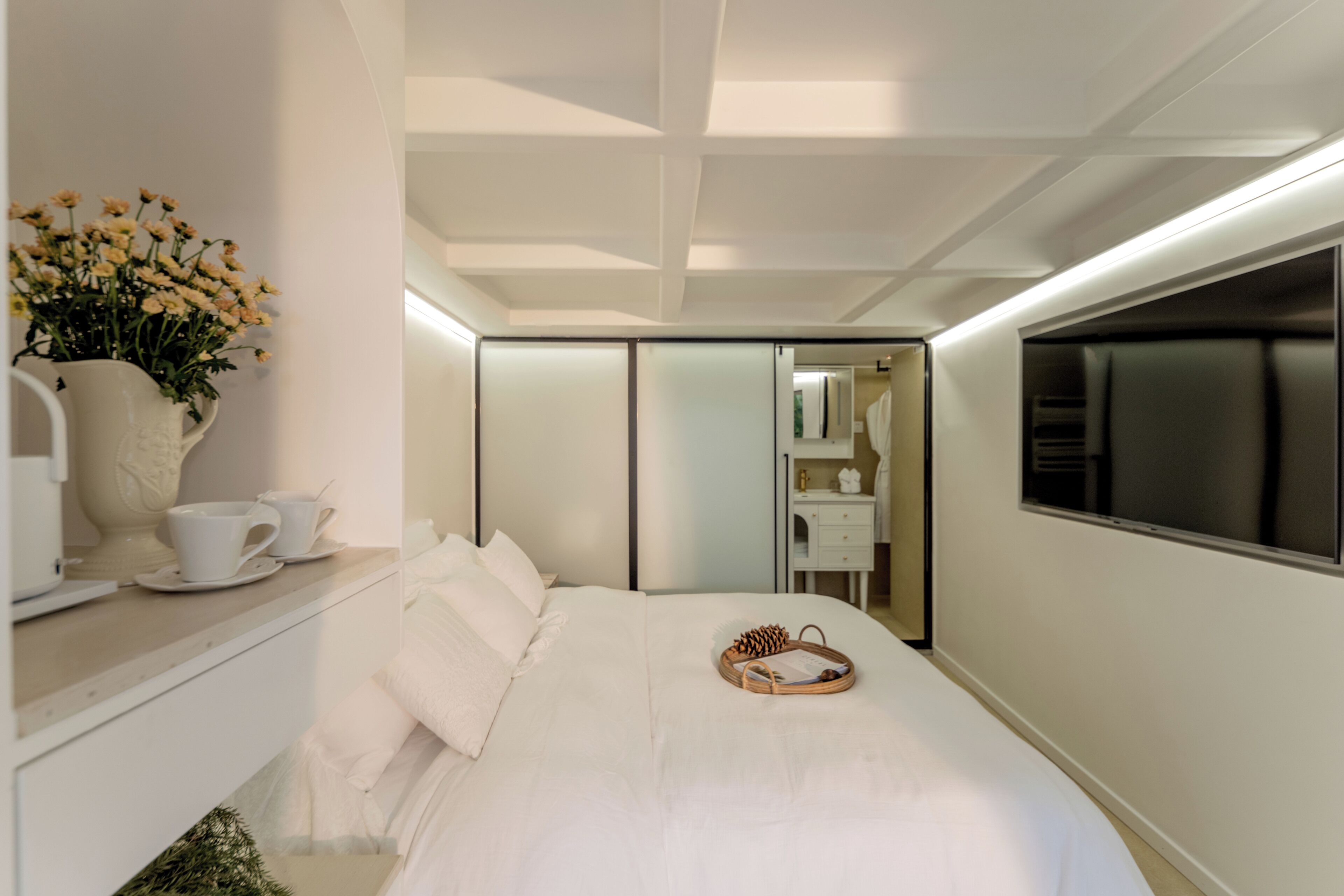 A modern hotel room with a neatly made bed, a bedside table with a vase of flowers, and a large mirror reflecting the room.