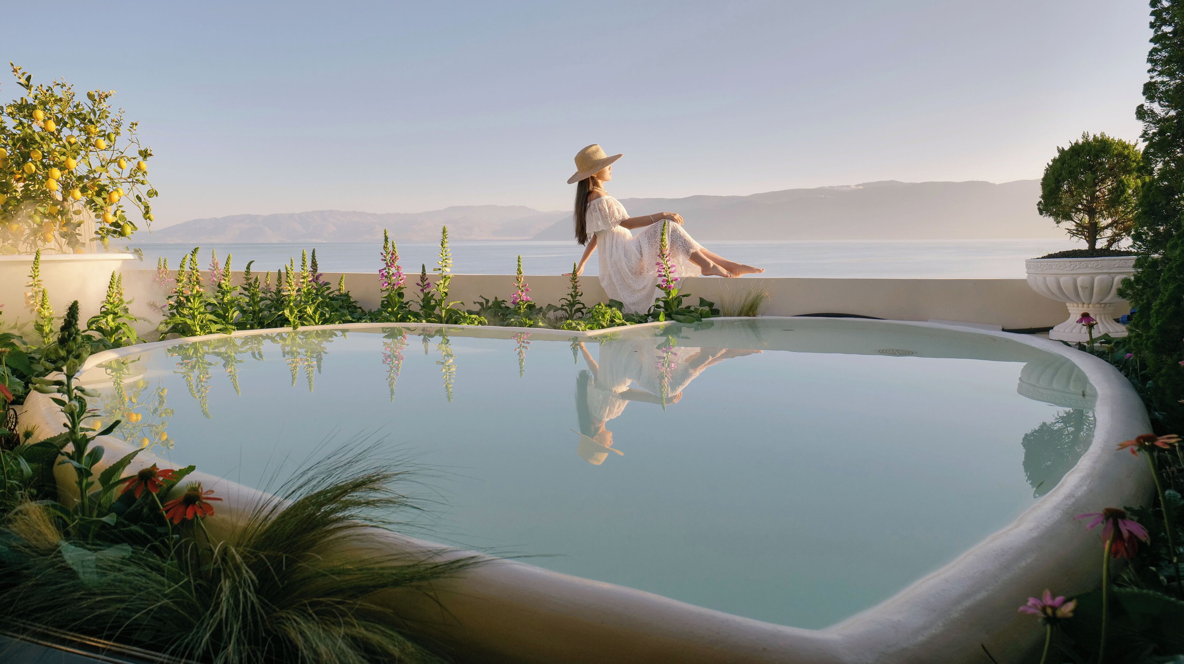 A woman sitting by a pool with a view of the sea and mountains.