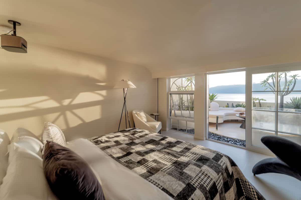 A bedroom with a large bed, a floor lamp, and a view of an outdoor seating area through large windows.