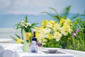 Una mesa dispuesta con una botella de vino, una copa y un florero con flores, con vistas al mar y follaje en el fondo.