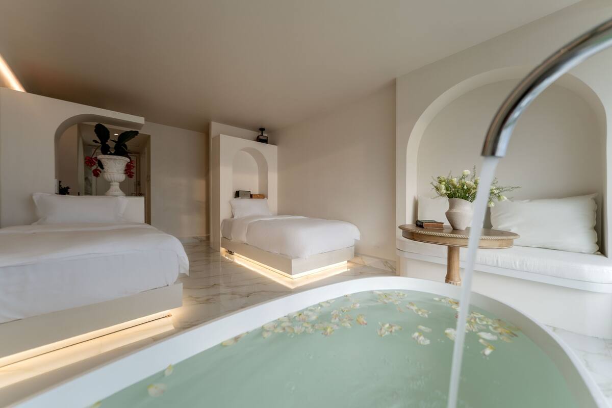 A modern hotel room with a large bathtub, a bed, and a small table with a vase of flowers.