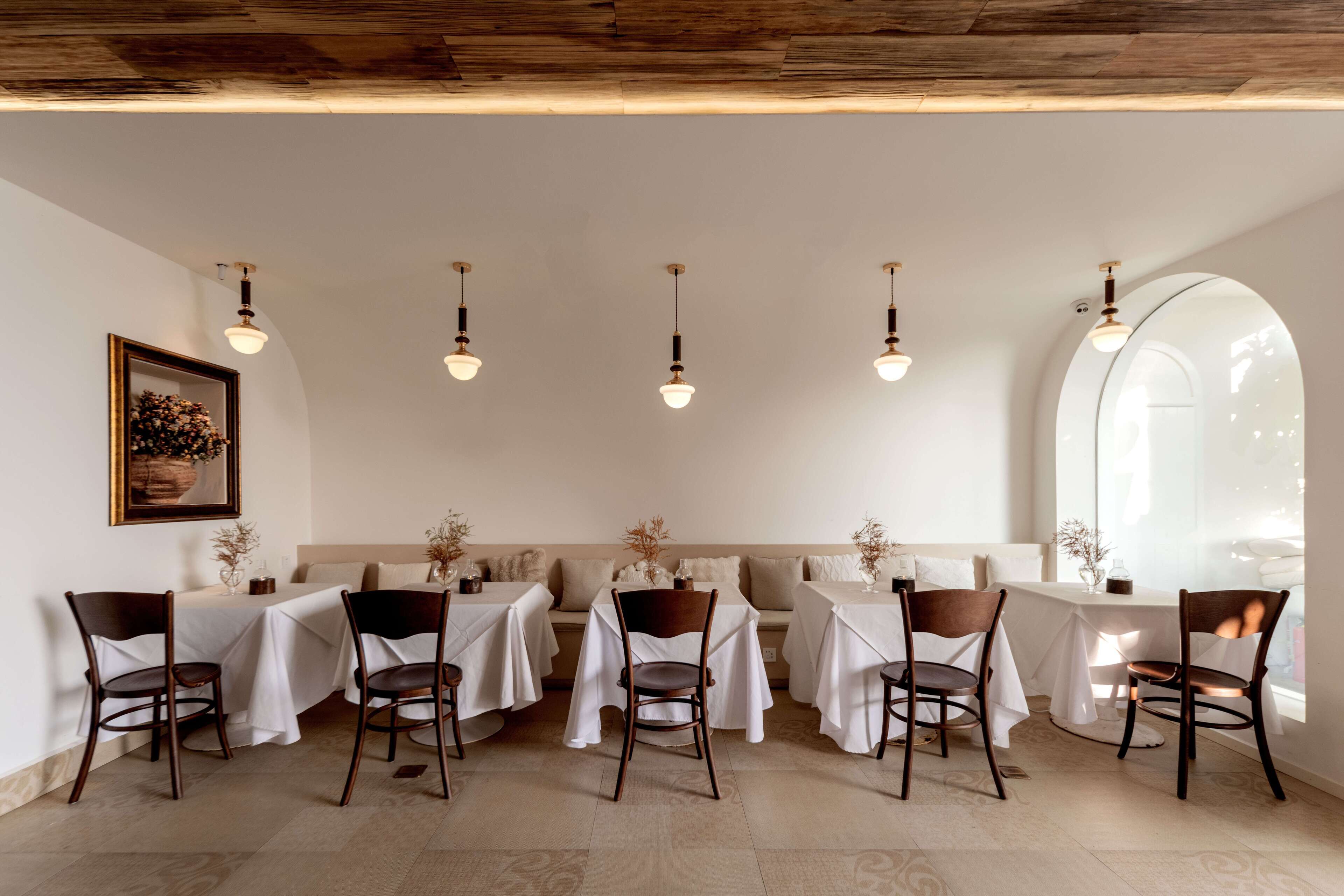 Un comedor con mesas dispuestas para seis personas, cada una con mantel blanco y sillas de madera. Hay lámparas colgantes sobre las mesas y un amplio ventanal arqueado a la derecha.