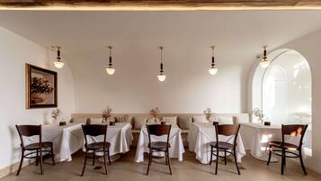 Un comedor con mesas dispuestas para seis personas, cada una con mantel blanco y sillas de madera. Hay lámparas colgantes sobre las mesas y un amplio ventanal arqueado a la derecha.