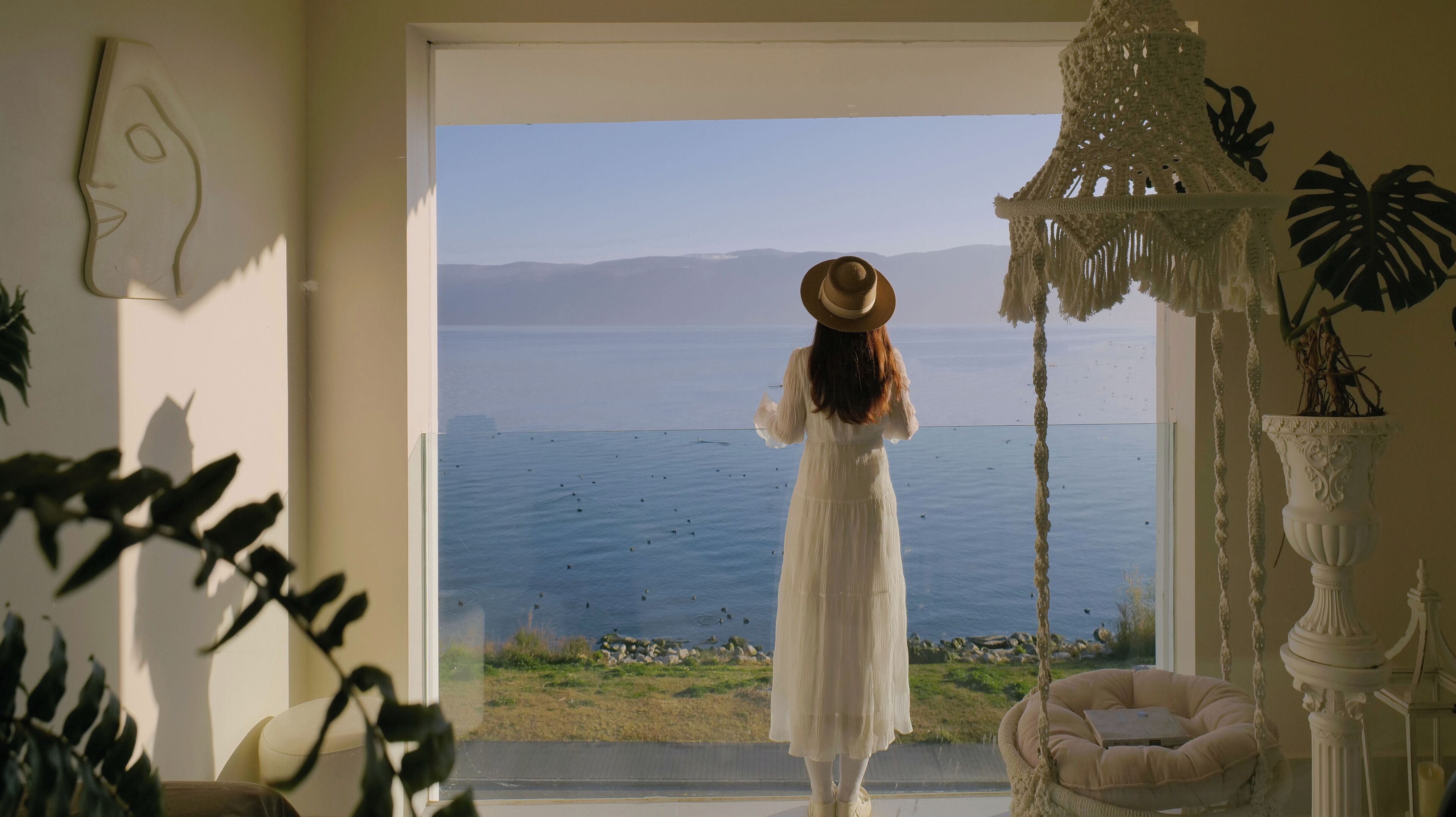 Una habitación con un ventanal amplio que da a una masa de agua, una silla blanca y una lámpara colgante de macramé.