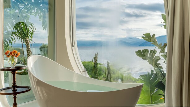 A modern bathroom with a freestanding bathtub, a small table with a vase of flowers, and a large window offering a view of a lake and mountains. - Dali Nest Secret Garden Lake View Inn (Dali)