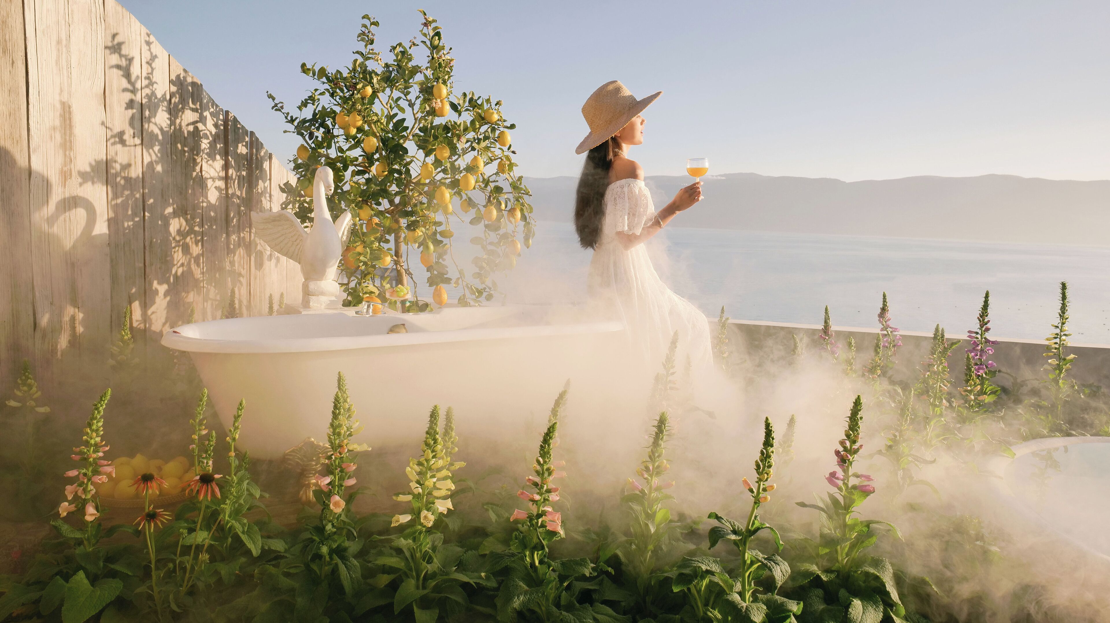 Une femme vêtue d’une robe blanche et d’un chapeau, près d’une baignoire, avec vue sur la mer et les montagnes.