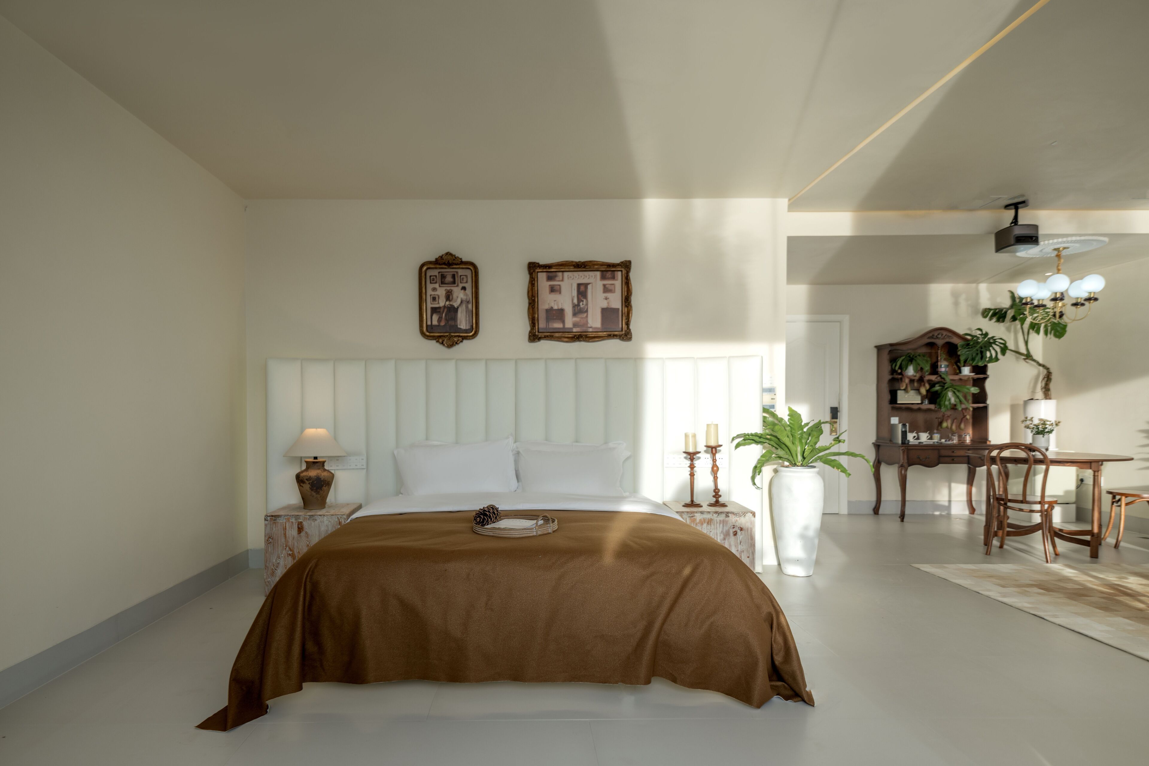 A bedroom with a large bed, a nightstand with a lamp, a dresser, and a dining area with a table and chairs.