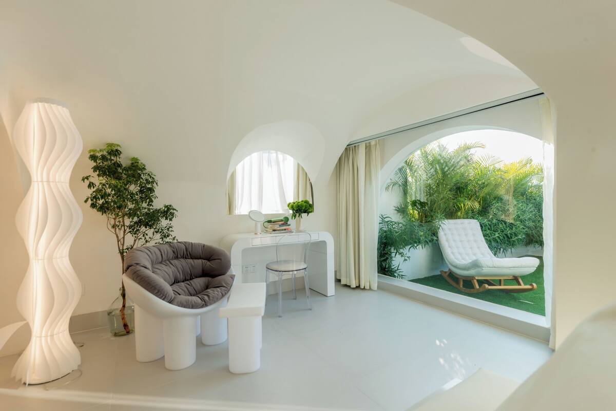 A modern interior with a curved ceiling, a white sofa, a dining table, and a rocking chair by a large window with a view of greenery.