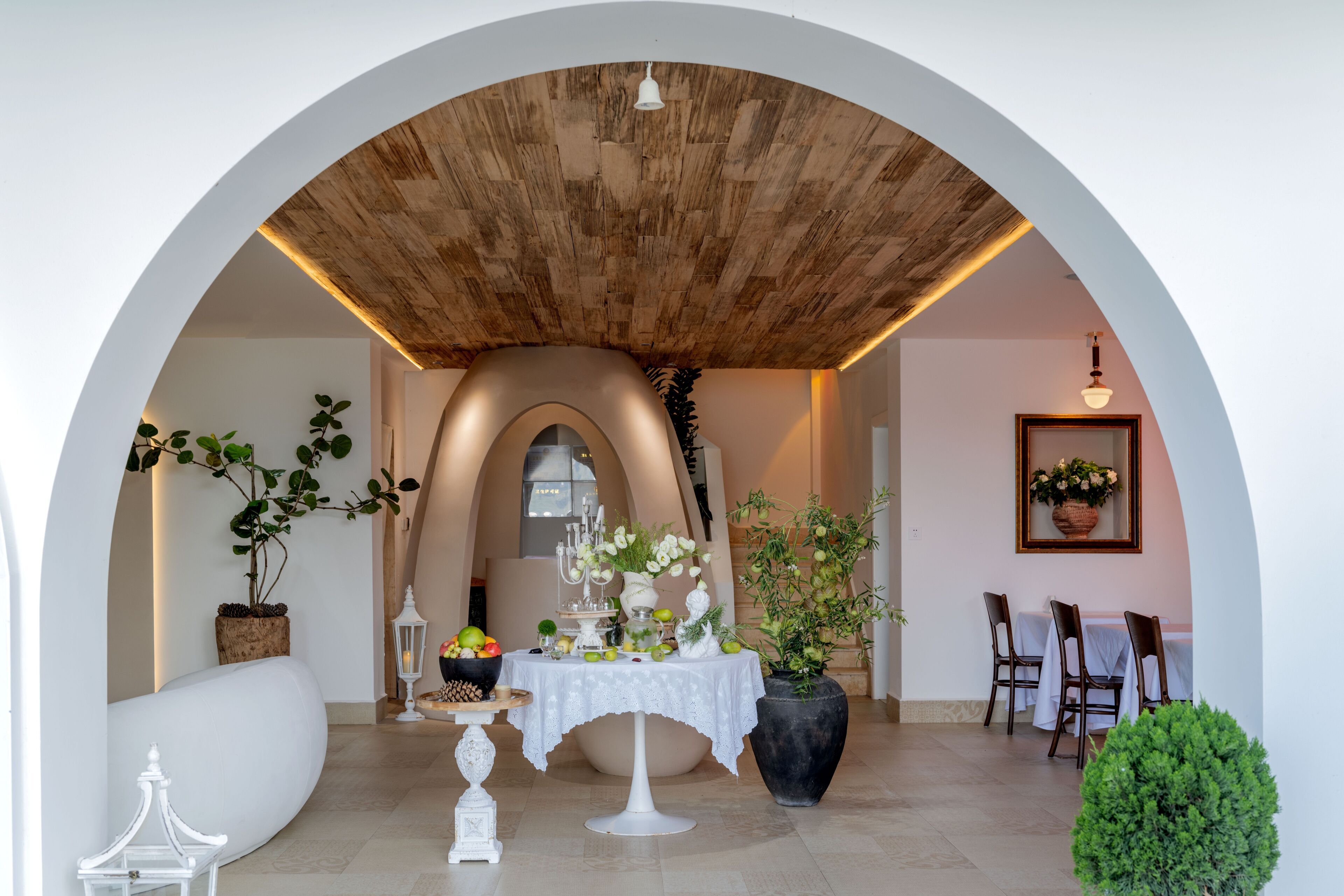 An indoor dining area with a white table set for a meal, surrounded by potted plants and a wooden ceiling.