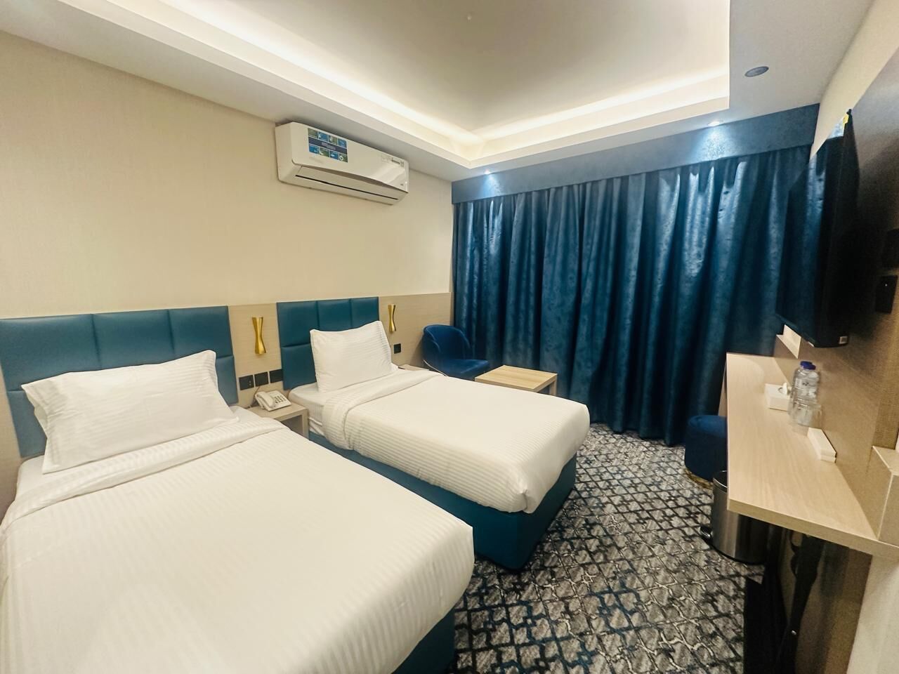 A hotel room with two beds, a desk, a chair, a TV, and an air conditioning unit.