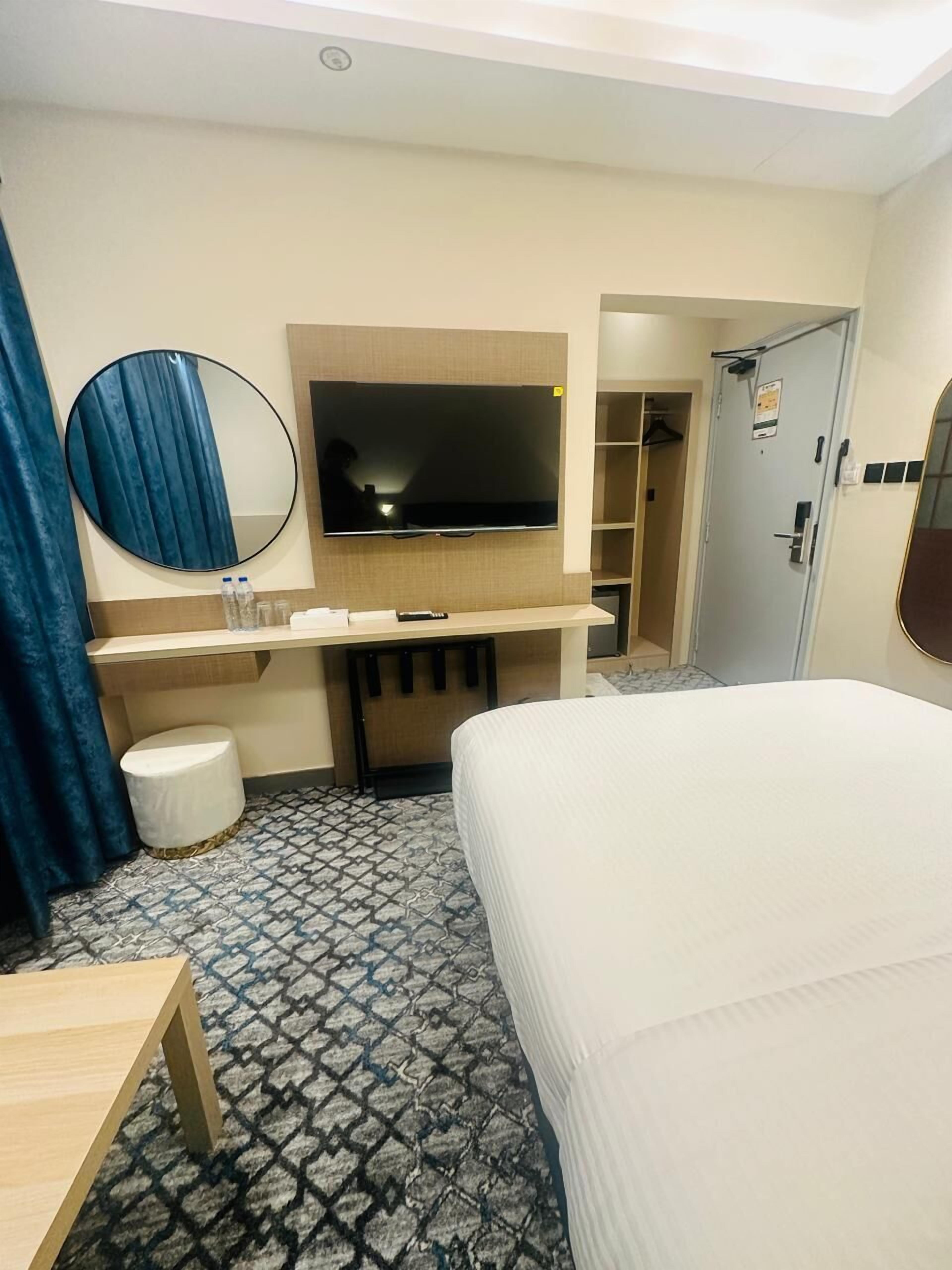 A hotel room with a bed, a TV mounted on the wall, a small desk, a mirror, and a bathroom visible through an open door.