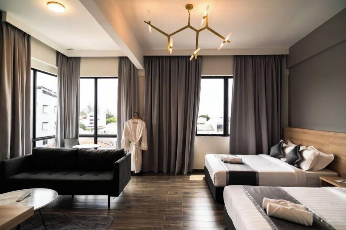 A modern hotel room with a sofa, two beds, a chandelier, and large windows.