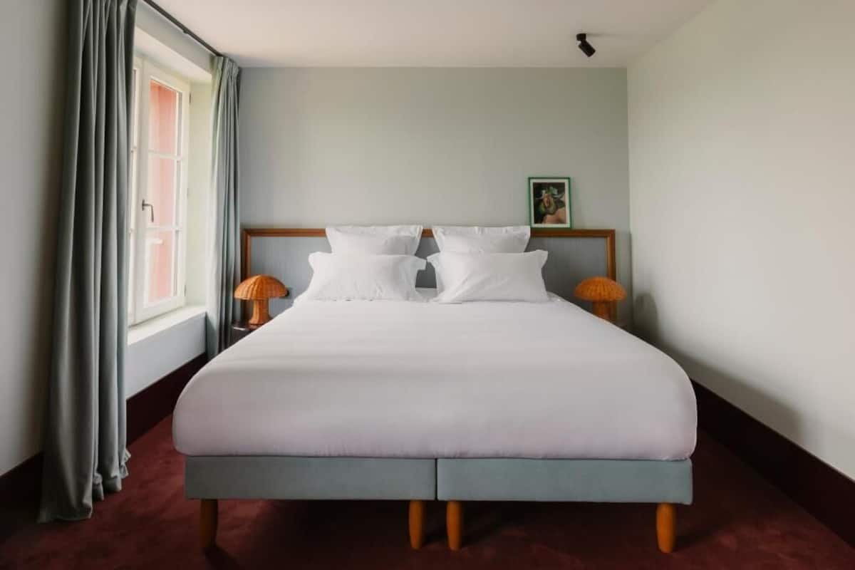 Una cama bien hecha con sábanas blancas, dos lámparas de noche y una ventana con cortinas.