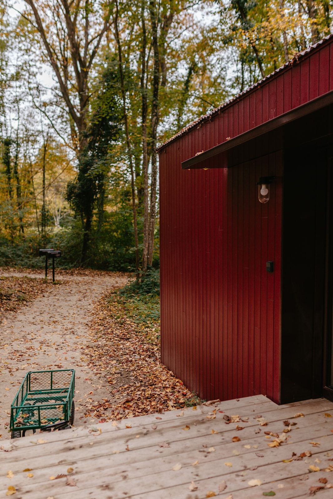 Un bâtiment rouge avec une porte et un luminaire, entouré d’arbres et d’une terrasse en bois parsemée de feuilles.