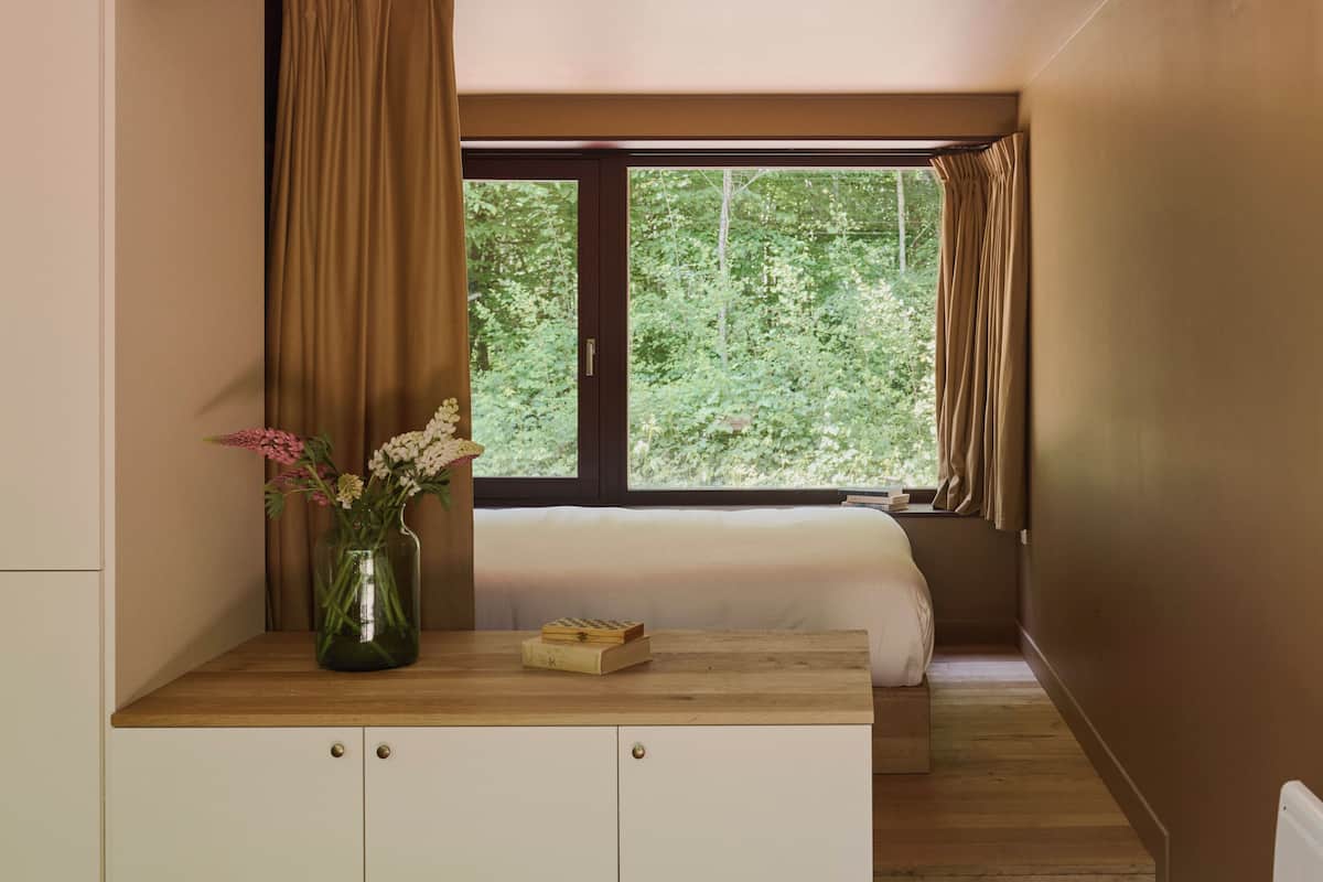 Un dormitorio con ventana, mesita de noche con un florero y una cama.